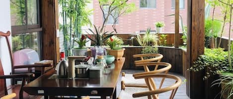 A wooden dining table with chairs, surrounded by potted plants, in a room with large windows and a view of a brick building.