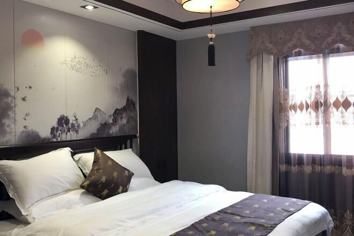 A hotel room with a large bed, a wall mural, a hanging light fixture, and a window with curtains.