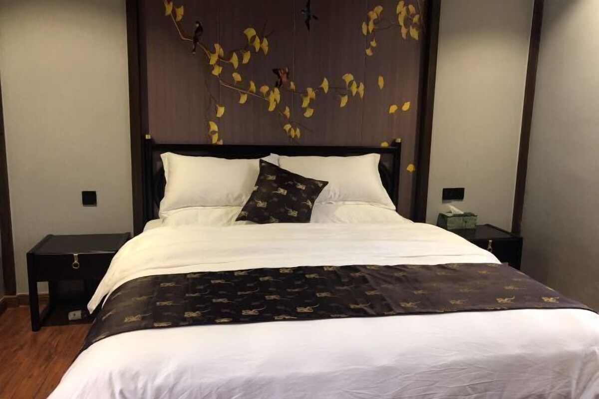 A neatly made bed with white linens and a dark patterned throw, a wooden headboard with leaf decorations, and a small bedside table with a lamp.
