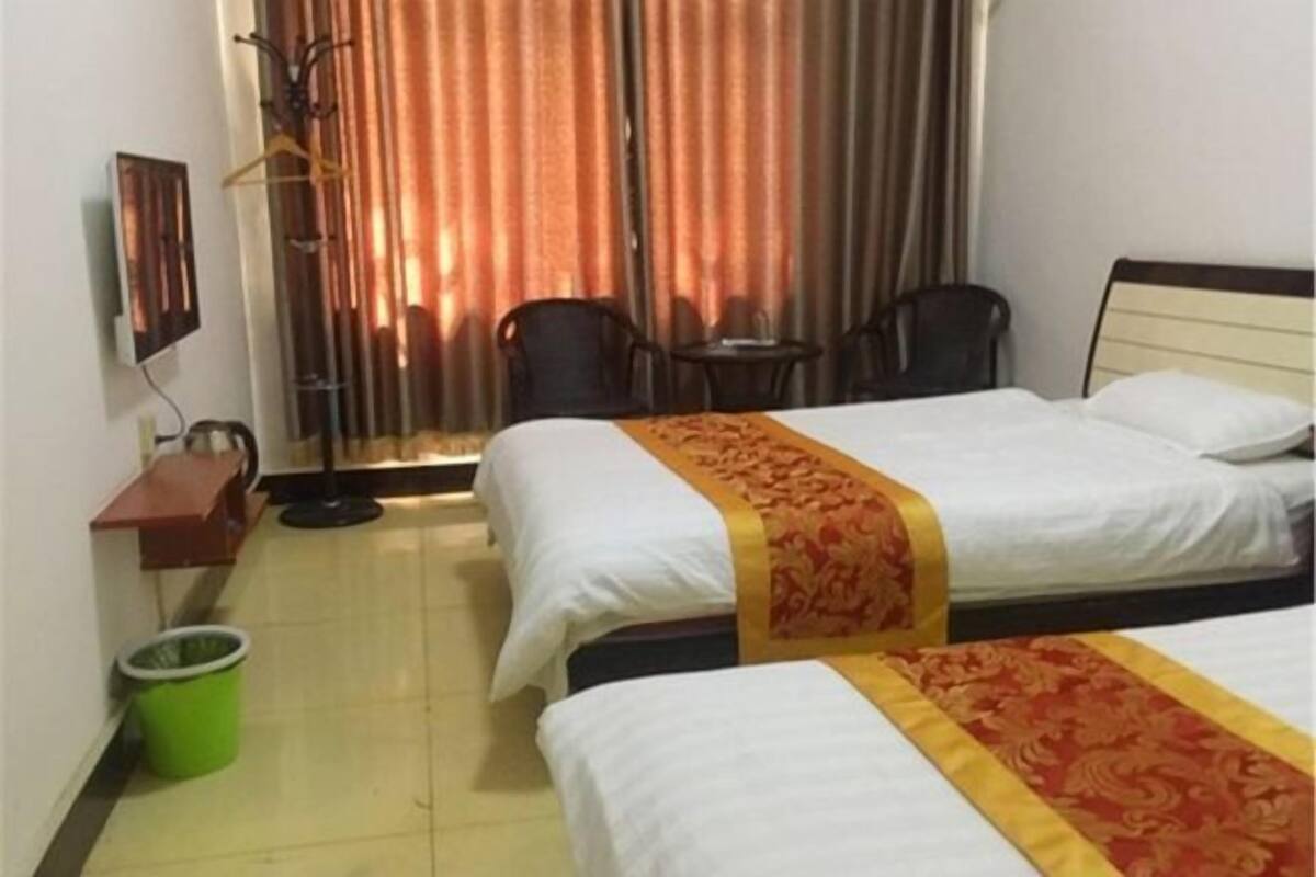Ein Hotelzimmer mit zwei Betten, einem kleinen Tisch, zwei Stühlen, einem Mülleimer und einem Fenster mit Vorhängen.