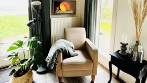 Ein Zimmer mit einem beigen Sessel, einer Blumentopf-Pflanze, einem gerahmten Kunstwerk, einer Kerze und einer Vase mit getrockneten Blumen.
