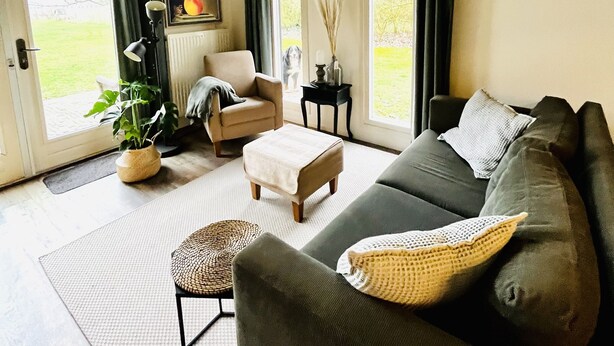 Ein modernes Wohnzimmer mit einem grauen Sofa, einem beigen Sessel, einem kleinen runden Hocker und einer Topfpflanze. Zwei Fenster mit Vorhängen, ein Heizkörper und ein gerahmtes Bild sind an der Wand zu sehen.