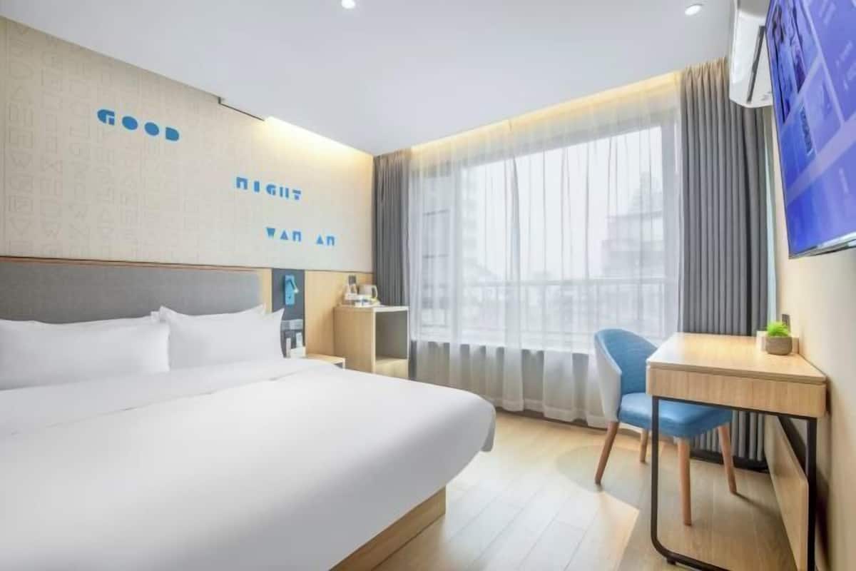 A hotel room with a large bed, a desk, and a chair. There is a window with curtains and a view of the city.
