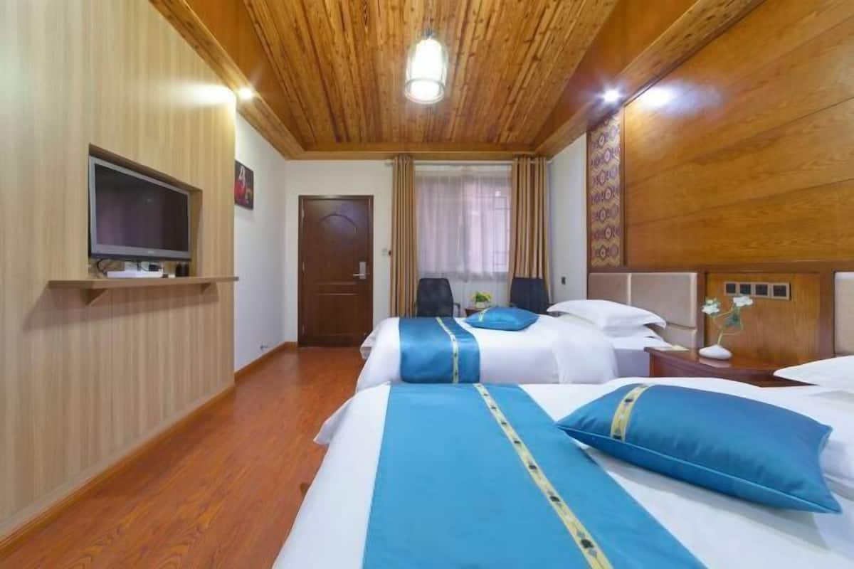 A hotel room with wooden paneling, a television, two beds with blue bedding, a window with curtains, and a wooden ceiling.