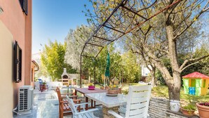 An outdoor patio with a stone table and white chairs, surrounded by trees and a small playhouse.