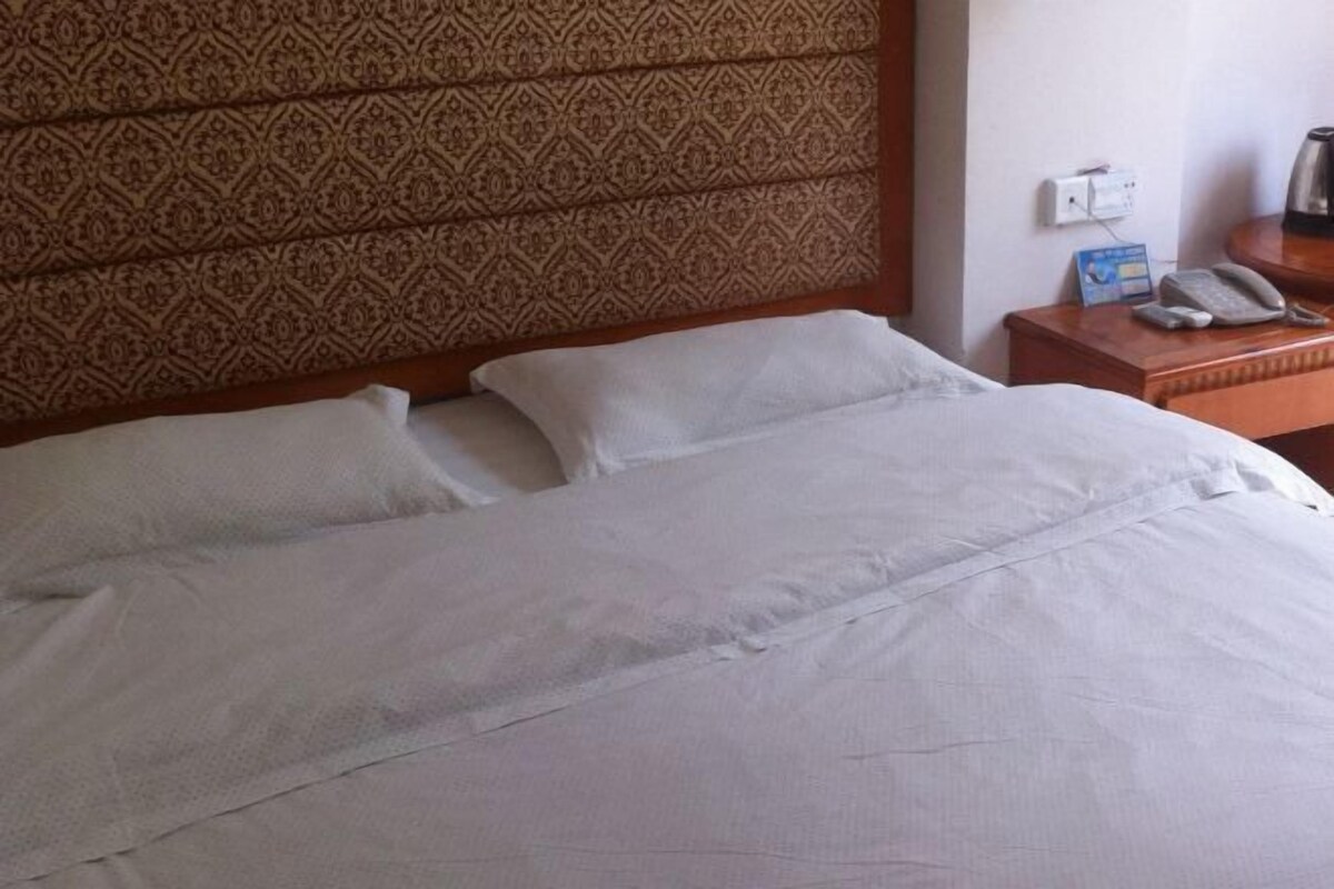 Une chambre d’hôtel avec une tête de lit à motifs, un lit bien fait avec du linge de lit blanc, une table de chevet avec un téléphone et une petite boîte, et une prise de courant au mur.