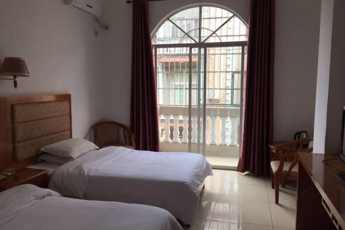 A hotel room with two beds, a wooden headboard, a desk with a chair, a TV, and a large window with curtains.