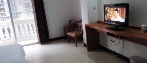 A hotel room with a bed, a TV on a wooden desk, a chair, and a view of a building through a window.