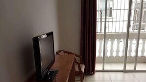 A room with a wooden desk, a chair, a television, and a view of a balcony with a railing.