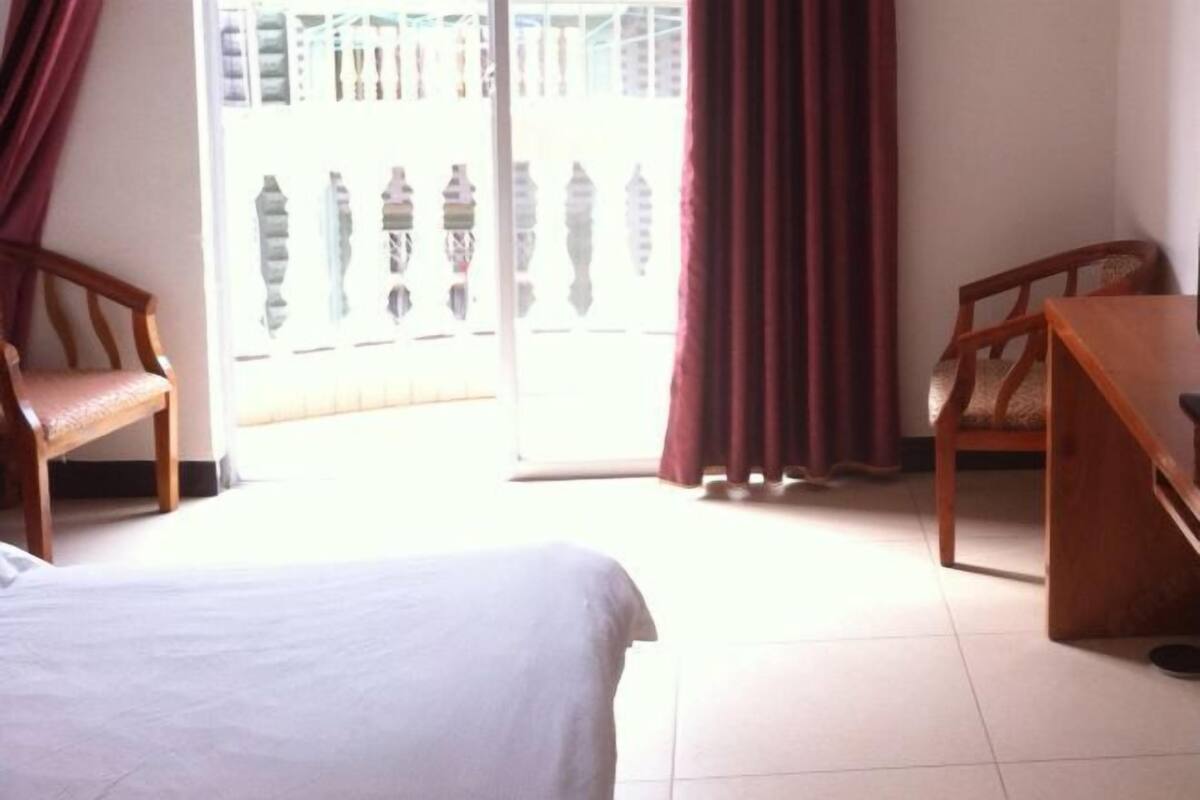 A hotel room with a bed, two wooden chairs, a window with a view of a building, and a sliding glass door.