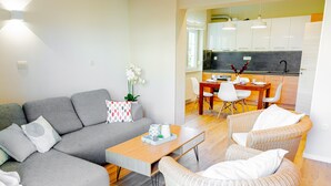 Ein modernes Wohnzimmer mit einem grauen Sofa, einem Holztisch und Korbstühlen. Im Hintergrund ist eine Küche mit weißen Schränken und einem Esstisch zu sehen.