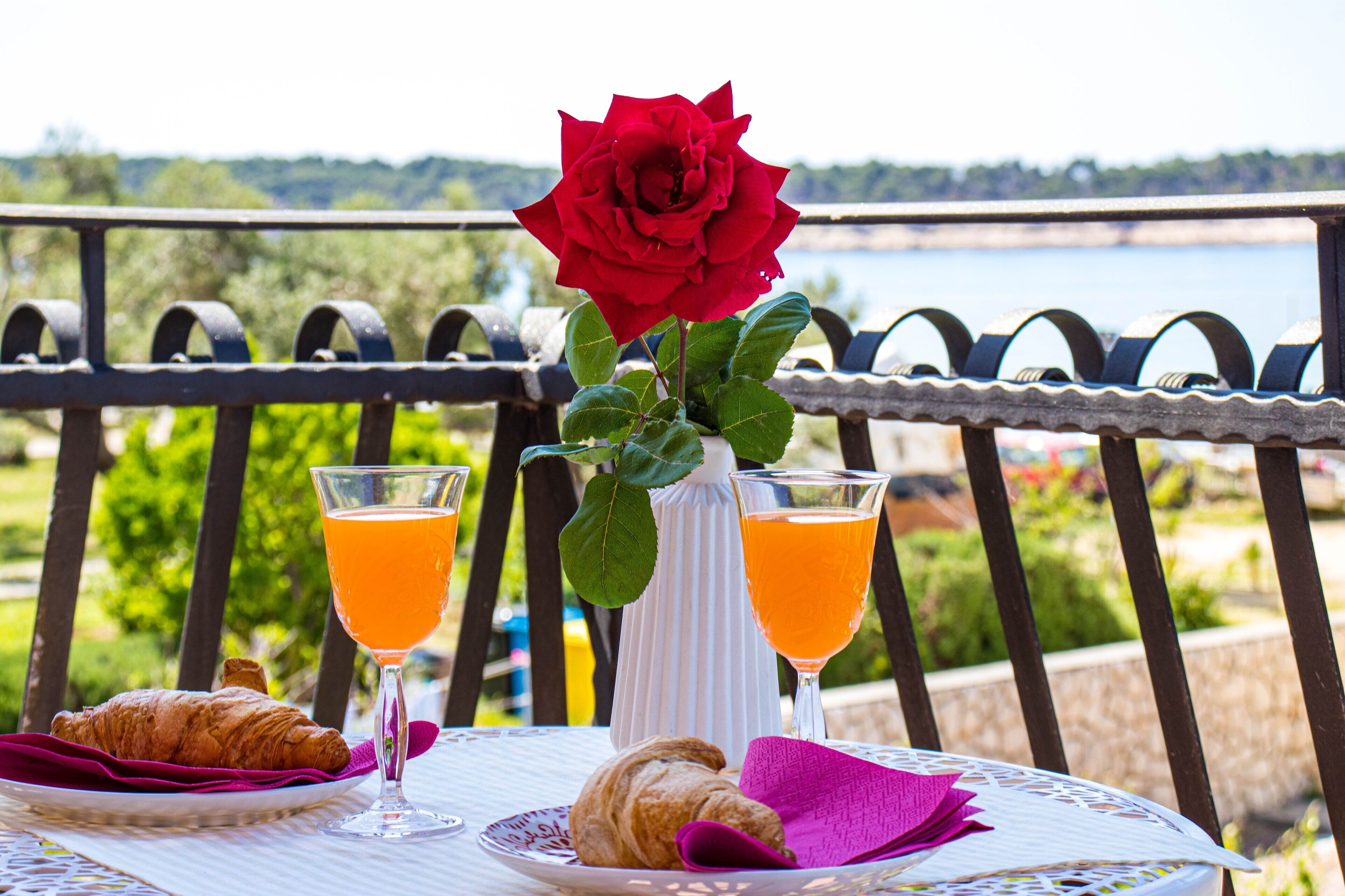 Uma mesa com croissants, dois copos de suco de laranja, um vaso com uma rosa vermelha e vista para um lago.