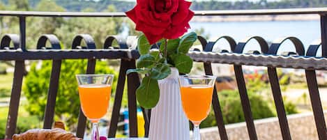 A table with croissants, two glasses of orange juice, a vase with a red rose, and a view of a lake.