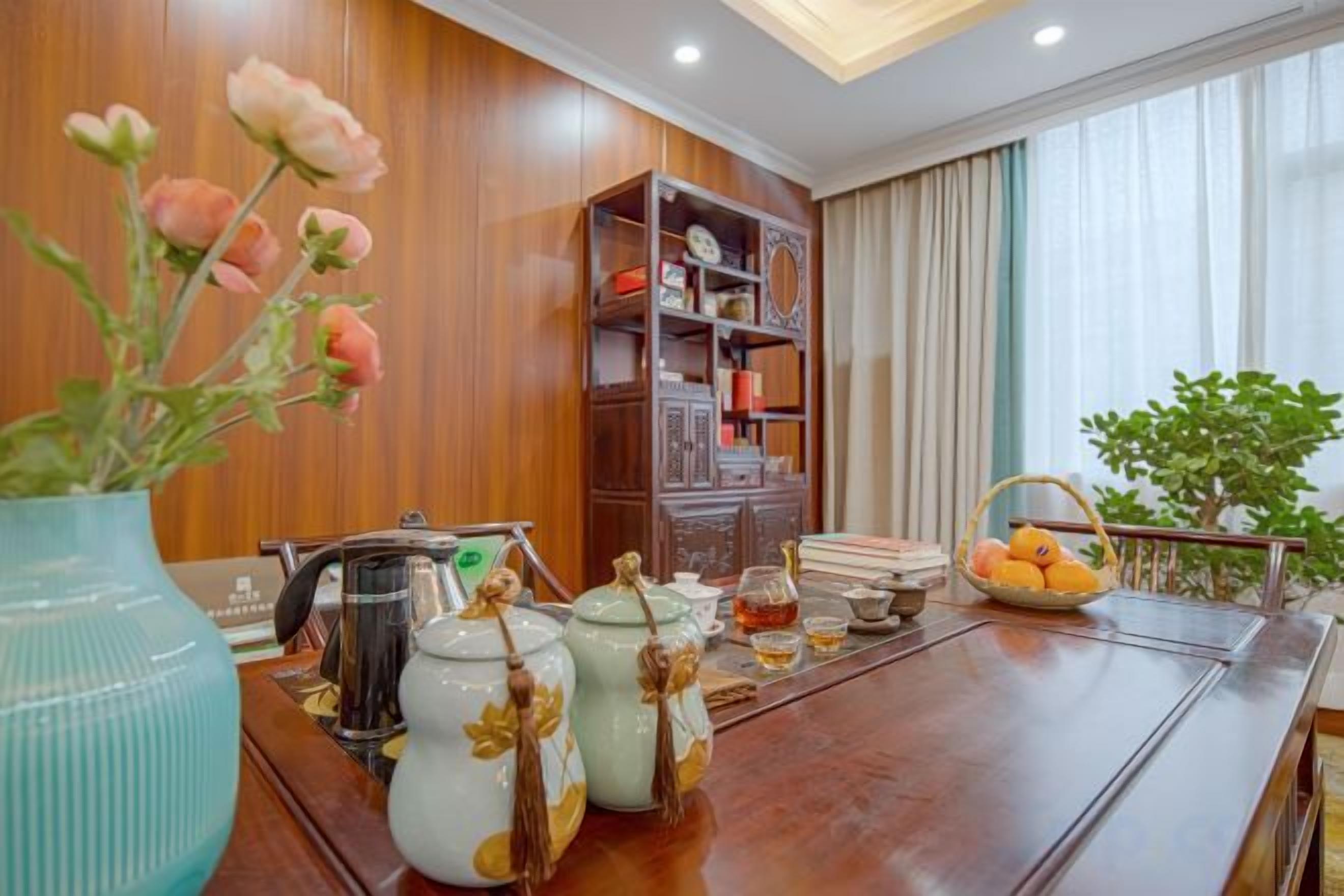 A dining table with tea sets, a vase with flowers, a bookshelf, and a window with curtains.