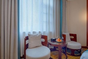 Habitación de hotel con una mesa redonda de madera, dos sillas, una alfombra pequeña y una ventana con cortinas de gasa.