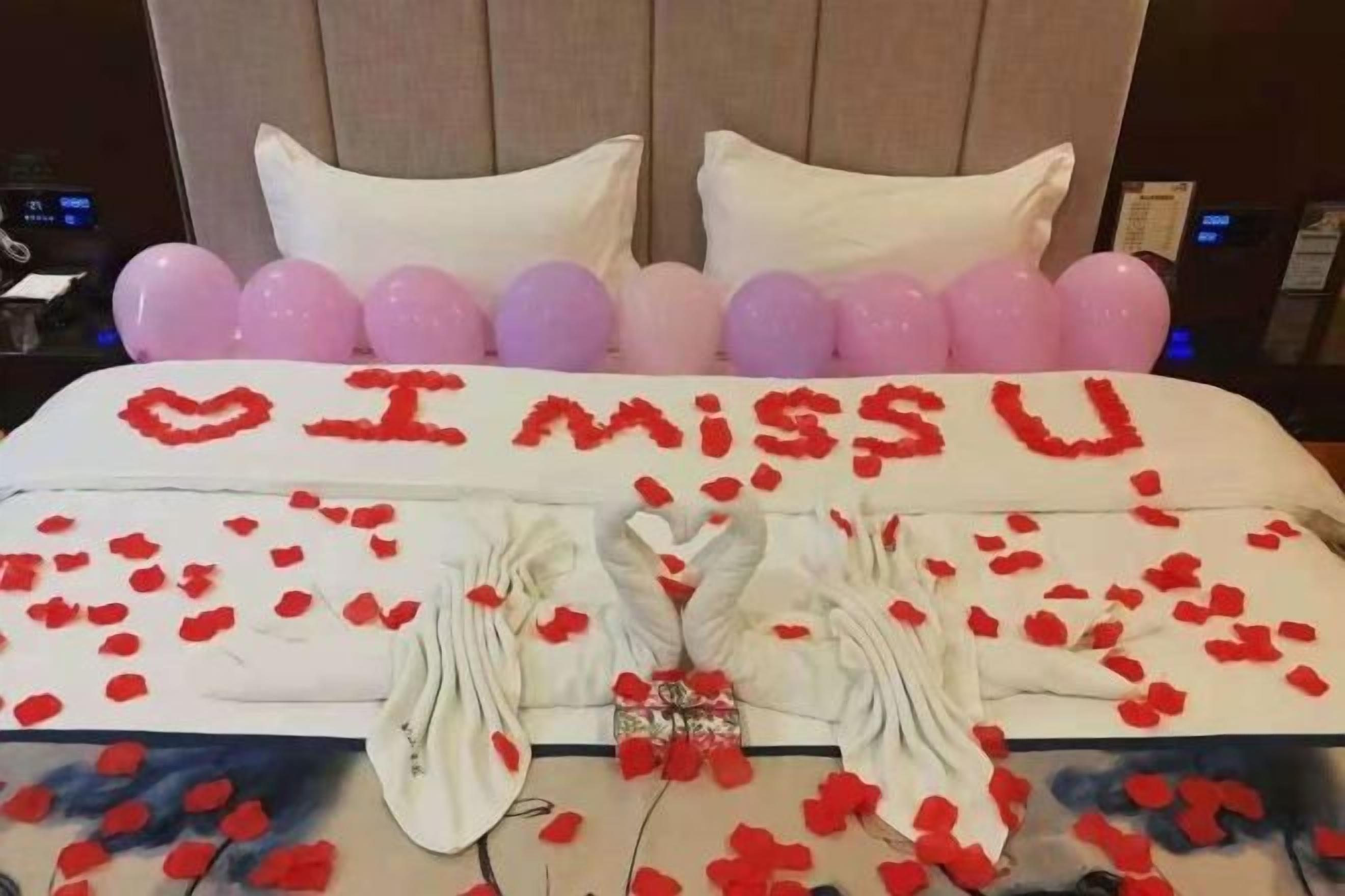 A bed with a heart-shaped arrangement of red rose petals spelling 'I miss you' and a bouquet of red roses.