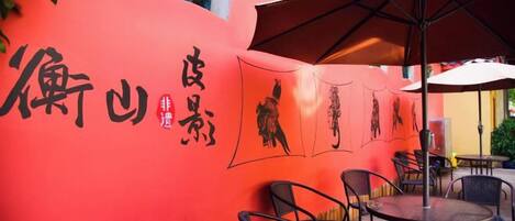 An outdoor seating area with tables and chairs, a red wall with black calligraphy, and a large umbrella providing shade.
