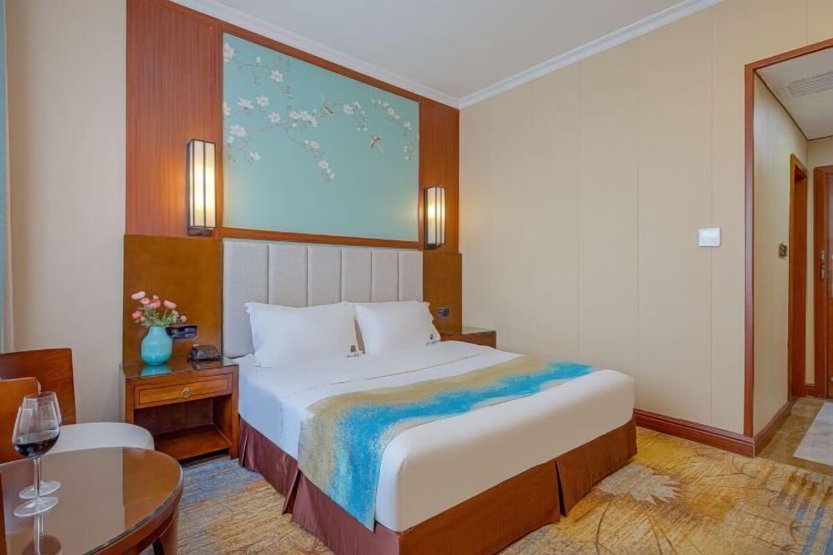 Una habitación de hotel con una cama grande, una mesita de noche, una mesa con una copa de vino y un adorno en la pared.