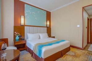 Una habitación de hotel con una cama grande, una mesita de noche, una mesa con una copa de vino y un adorno en la pared.
