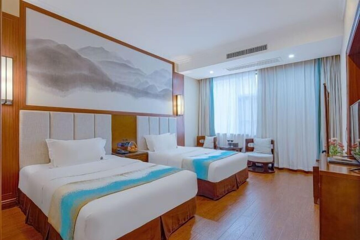 Habitación de hotel con dos camas, un cuadro grande en la pared, un escritorio con una silla y una ventana con cortinas.