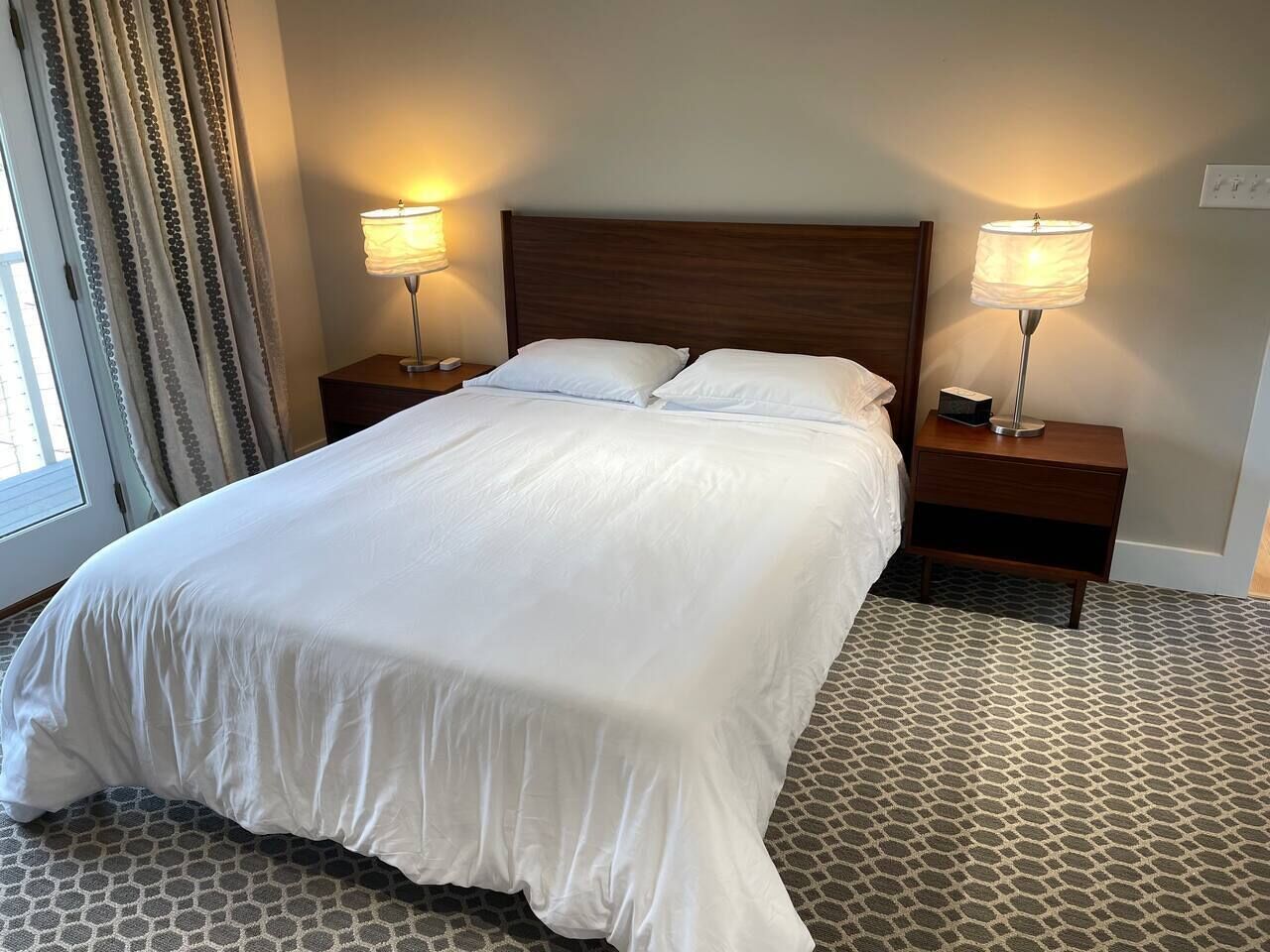 Ett hotellrum med en stor säng, två sängbord med lampor, en mönstrad matta och ett fönster med gardiner.
