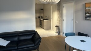 Una sala de estar moderna con un sofá de cuero negro, una mesa redonda blanca y una silla azul. Se aprecia una cocina al fondo con una isla blanca y una lámpara colgante.