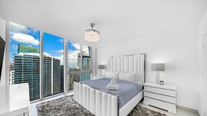 Une chambre moderne avec un grand lit, une table de chevet blanche, une lampe et une vue sur les gratte-ciel de la ville, visible à travers de grandes fenêtres.