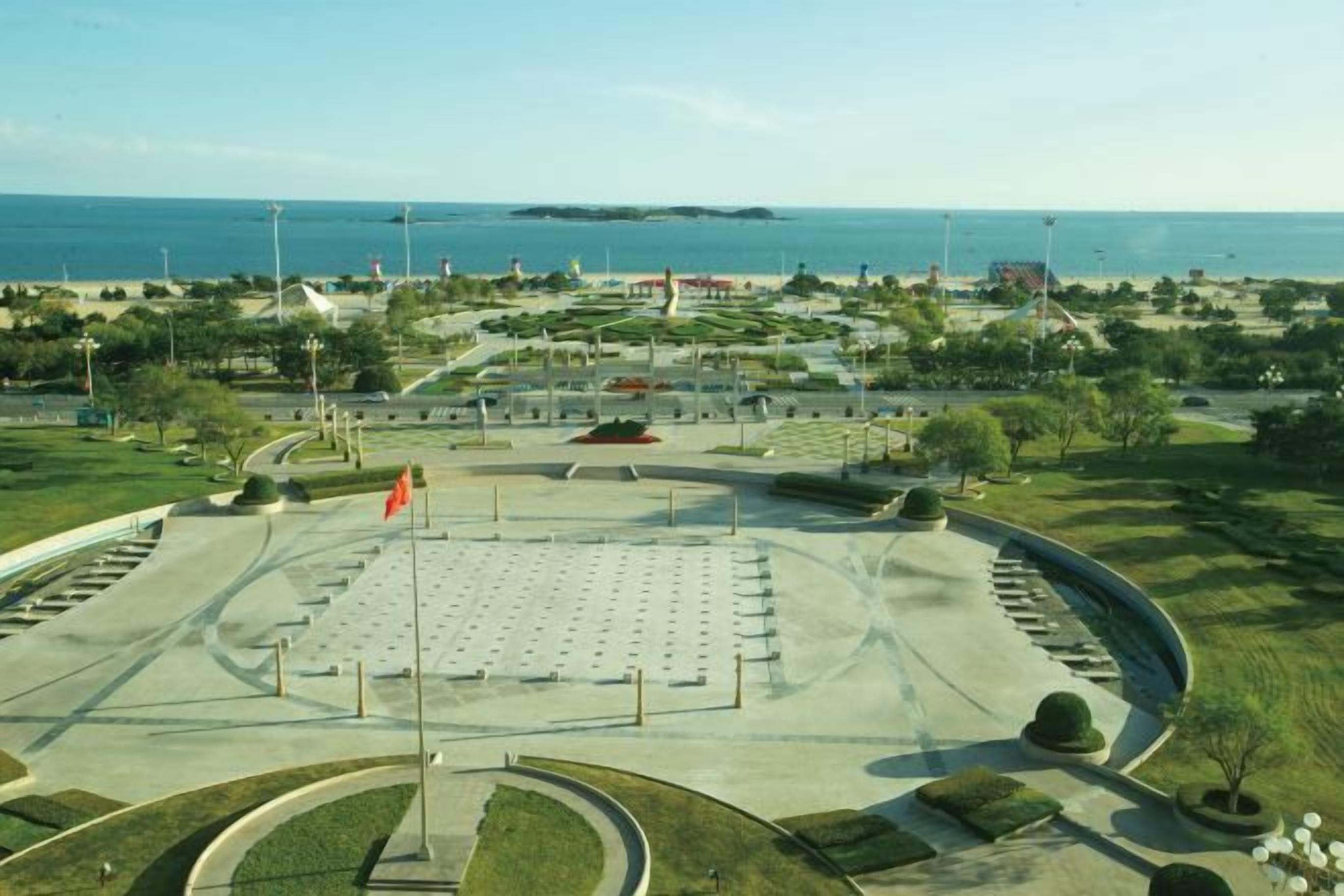 Une vaste place publique, avec un monument central, entourée de verdure et offrant une vue sur la mer.