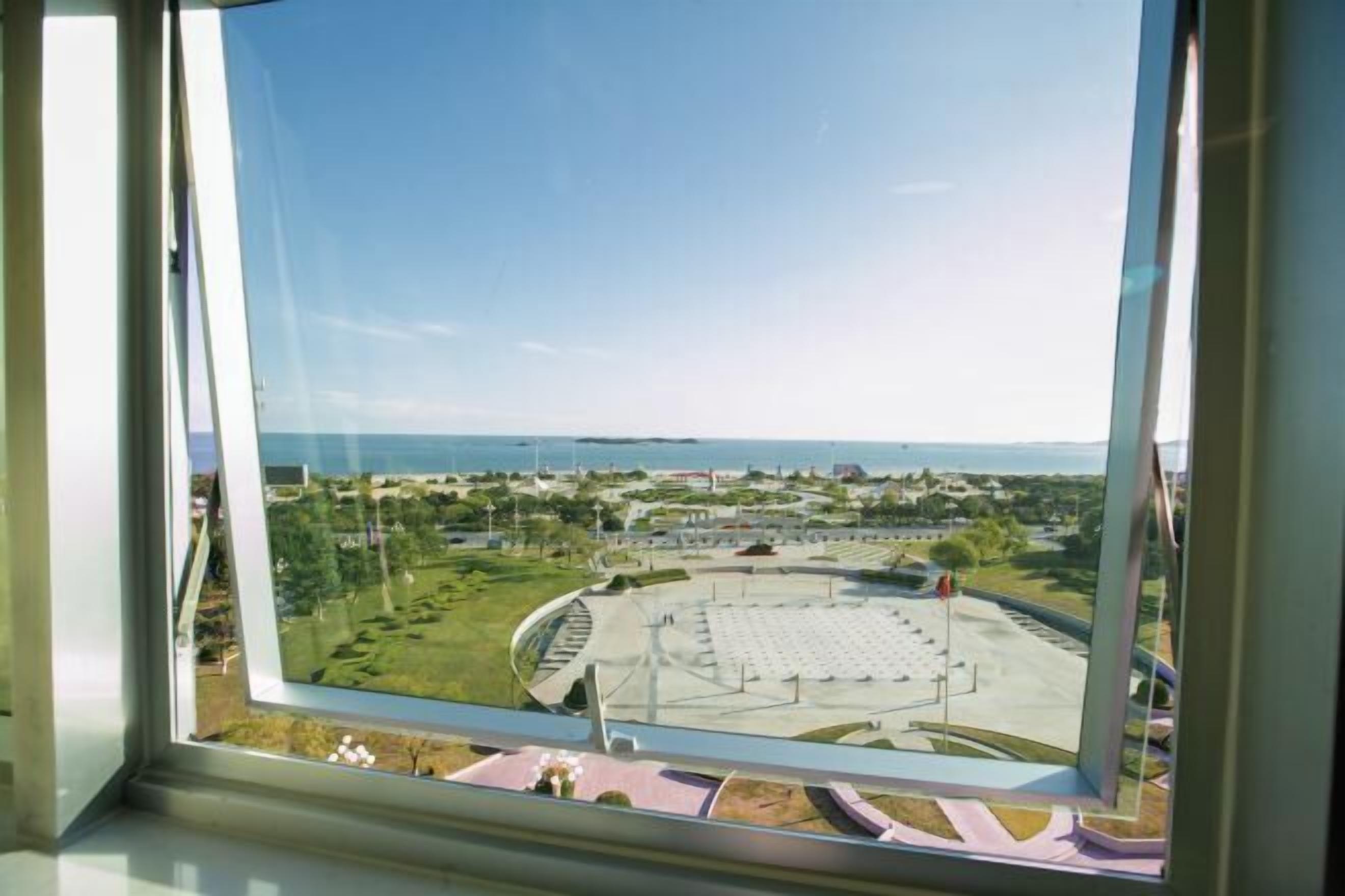Une vue depuis une fenêtre donnant sur une vaste place ouverte, avec un espace de repos circulaire et une étendue d'eau en arrière-plan.