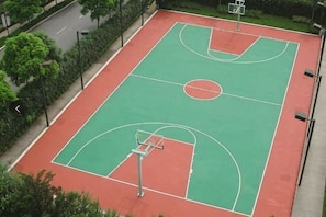 Uma quadra de basquete externa com superfície vermelha e verde, cercada por árvores e uma estrada.