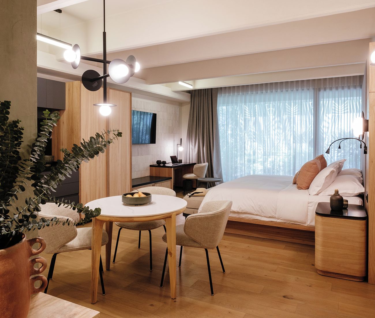 Una habitación de hotel moderna con cama, mesita de noche, lámpara y un área de comedor con sillas y mesa.