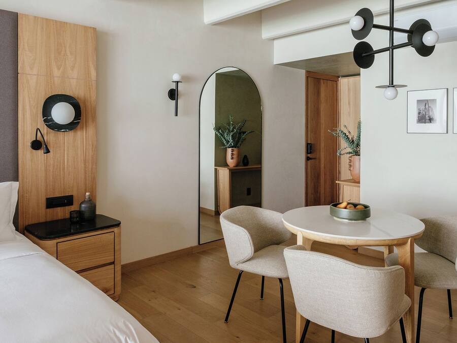 Une chambre d’hôtel moderne comprenant un lit, une table de chevet, une lampe, une table de salle à manger ronde avec des chaises et un miroir.