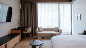 Ein modernes Hotelzimmer mit einem großen Bett, einer Couch, einem kleinen Tisch, einem Fernseher und einem Fenster mit Vorhängen.