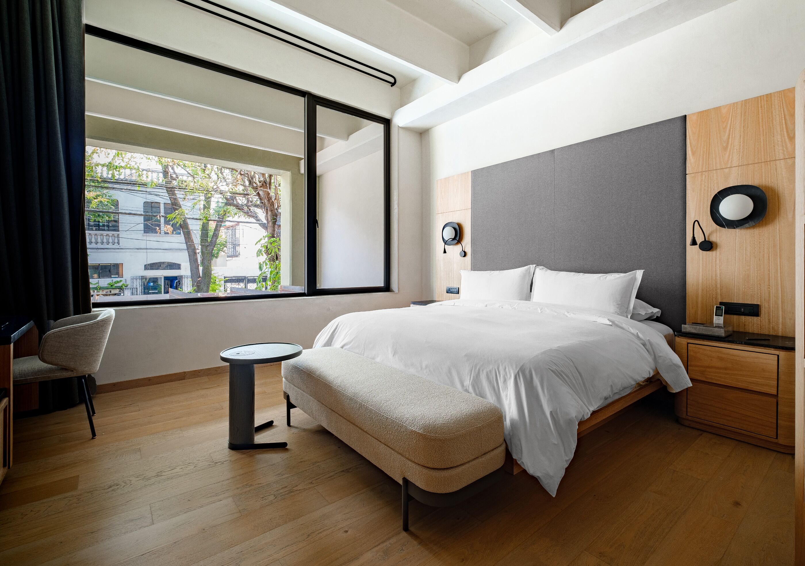 A modern bedroom with a large bed, a small round table, a chair, and a view of the outside through a large window.