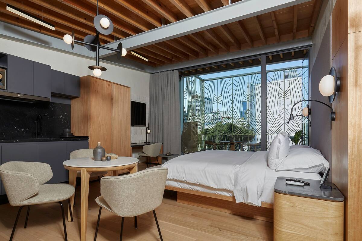 Una habitación de hotel moderna con techo de madera, una cama grande, un escritorio con silla, una mesita y una ventana con vistas a edificios.