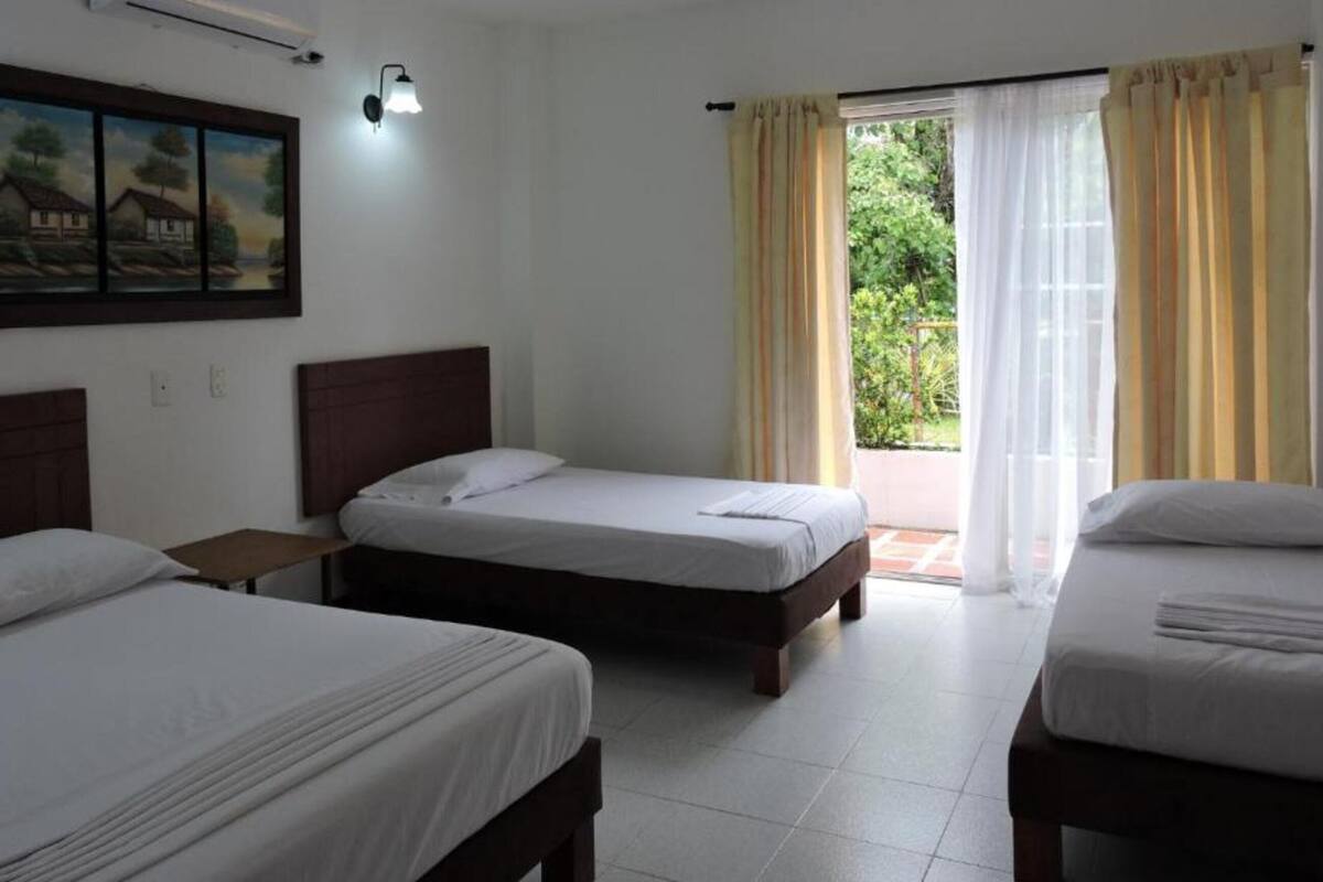 A hotel room with three single beds, a painting on the wall, a window with curtains, and a tiled floor.