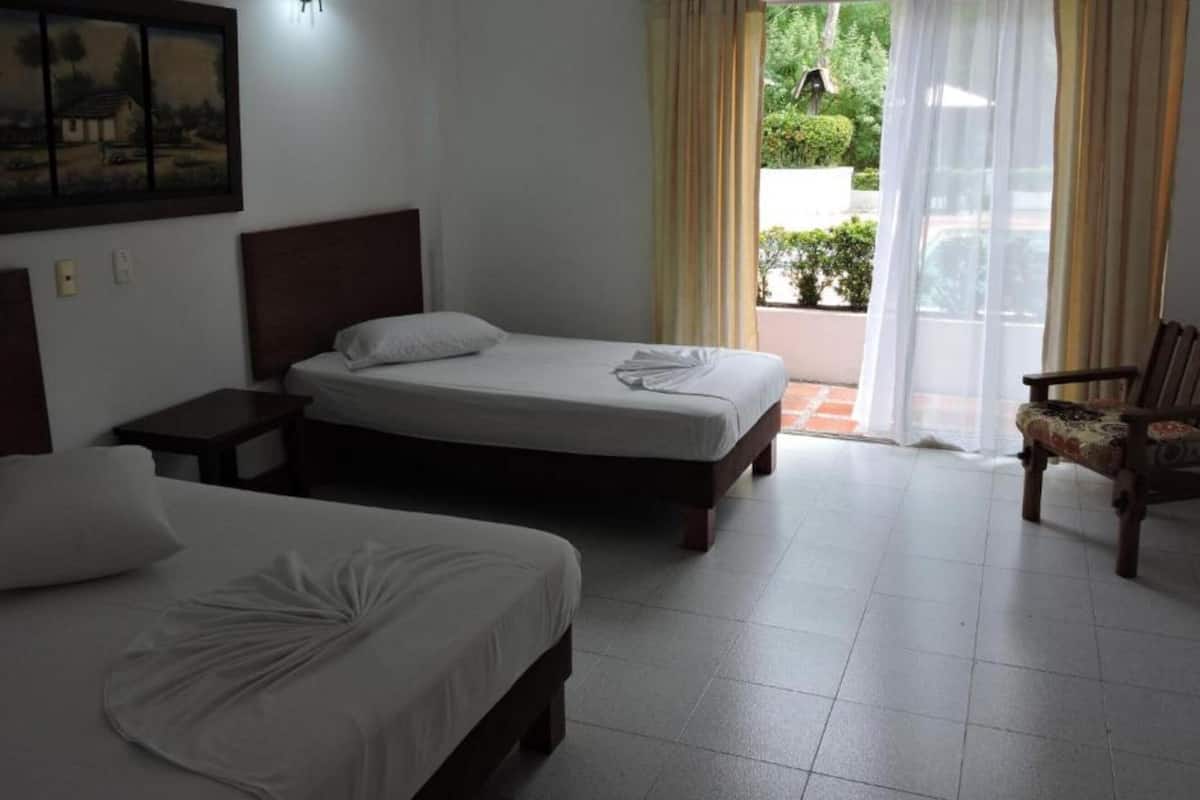 A hotel room with two beds, a wooden headboard, a chair, and a view of greenery through the window.