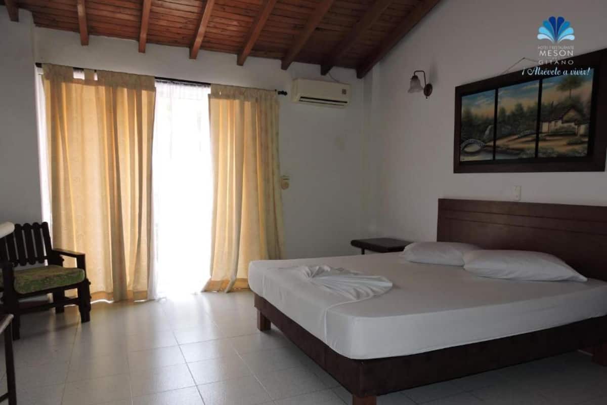 A hotel room with a bed, a bench, a painting on the wall, and a window with curtains.