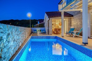 A swimming pool with a stone wall on one side, a covered patio area with seating, and a view of a building with a tiled roof. - Villa Belleza - sea views, heated pool, 70m to the beach by MyWaycation (Rogoznica)