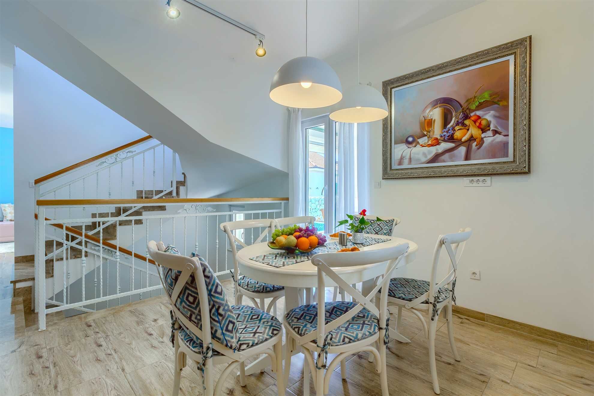 Un coin repas avec une table blanche, des chaises à motifs et un saladier rempli de fruits. On aperçoit un escalier au fond.