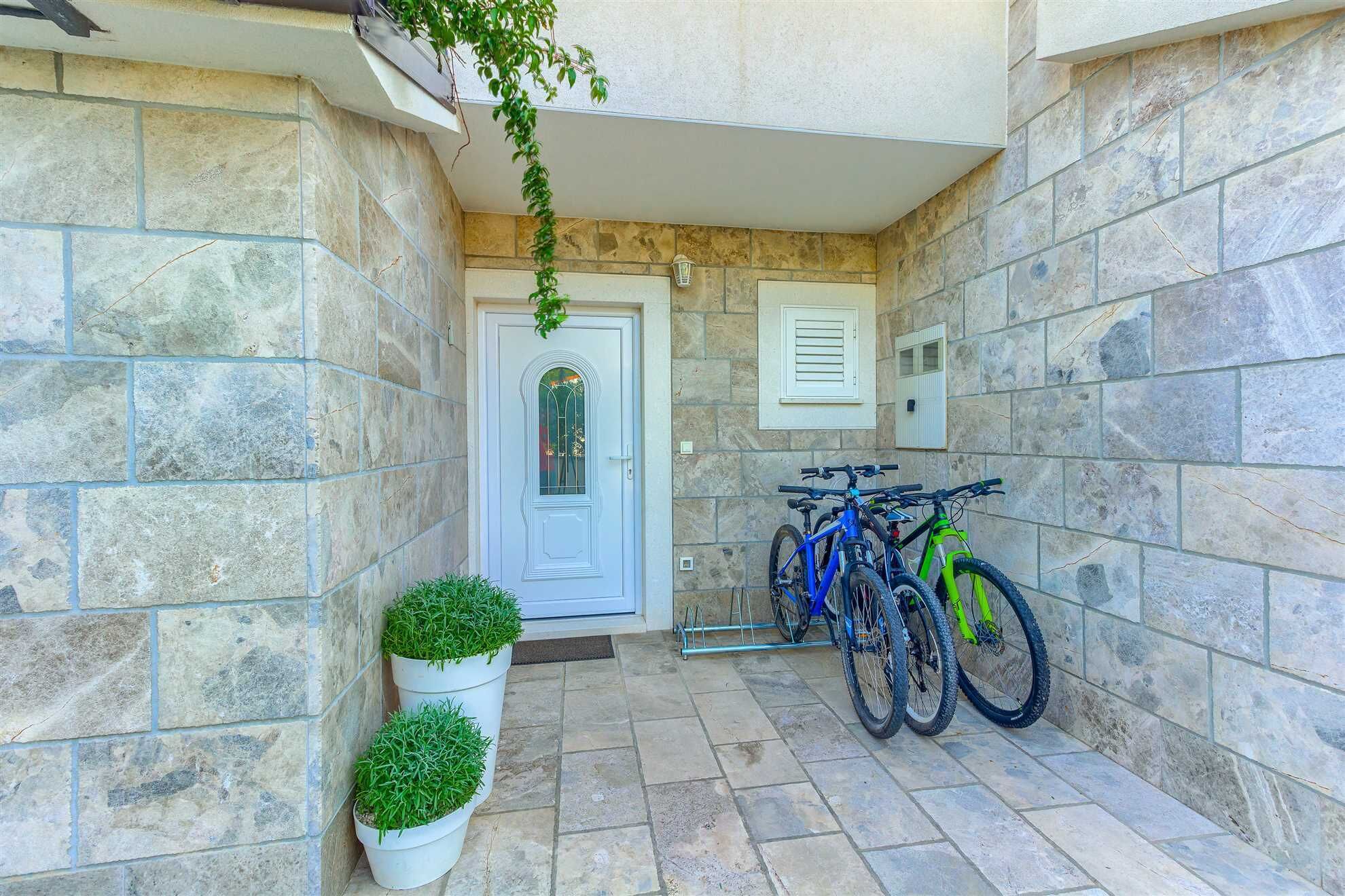 A stone-walled entrance with a white door, two potted plants, and two bicycles parked against the wall.