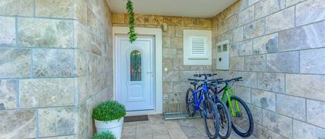 A stone-walled entrance with a white door, two potted plants, and two bicycles parked against the wall.
