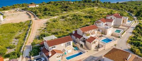 A coastal residential area with modern houses featuring red-tiled roofs, swimming pools, and a view of the sea.