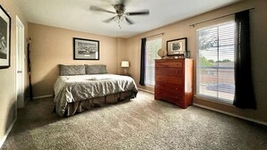Een slaapkamer met een bed, een dressoir, een plafondventilator, een raam met jaloezieën en een lijst met een afbeelding aan de muur.