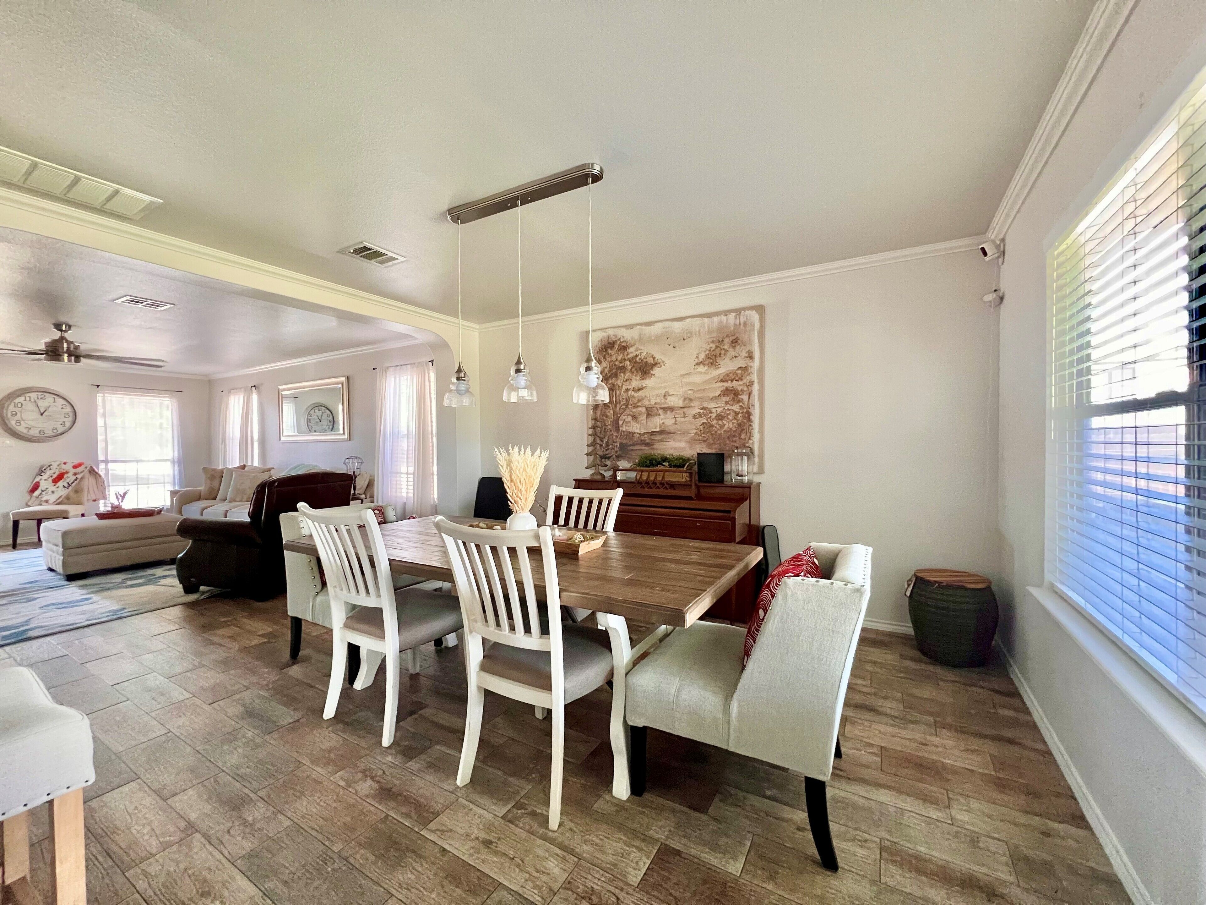 Un coin repas avec une table en bois et des chaises blanches, un tableau au mur et une fenêtre avec des stores.