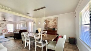 Een eetgedeelte met een houten tafel en witte stoelen, een schilderij aan de muur en een raam met zonwering.