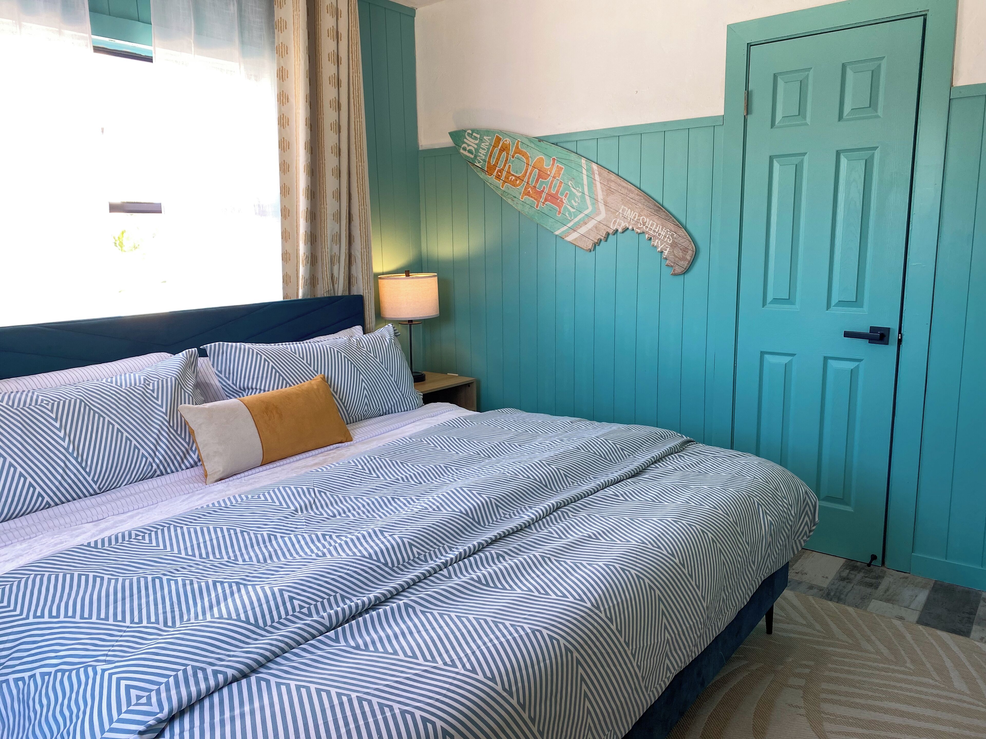 A bedroom with a bed, pillows, a nightstand, a lamp, a teal door, and a wall decoration.