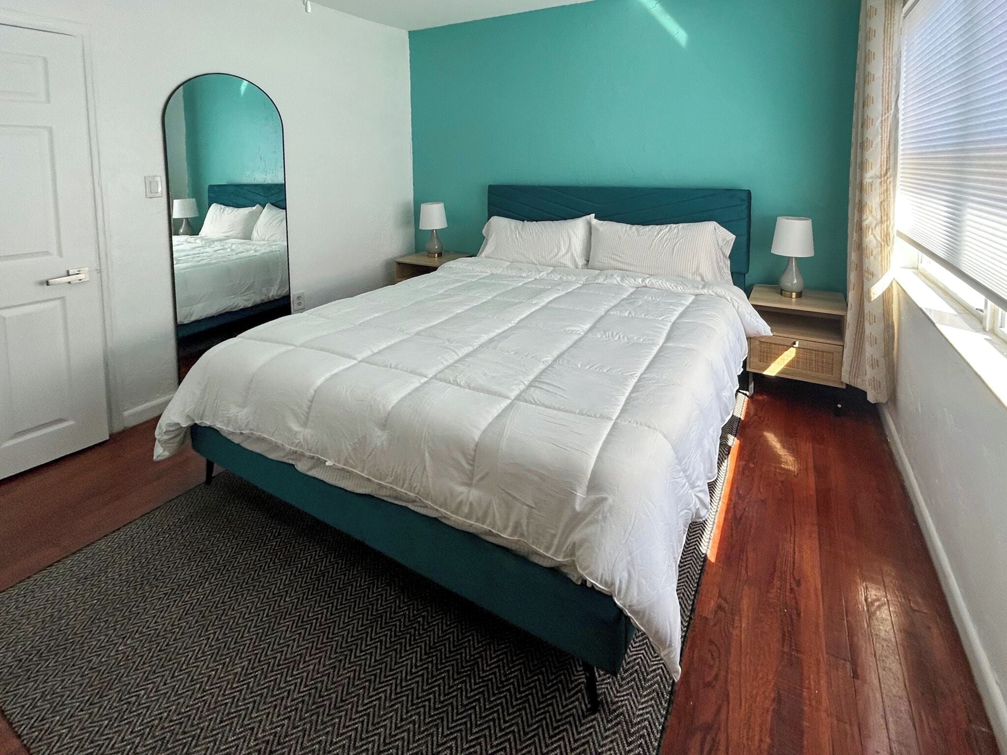 Une chambre avec un grand lit, un miroir, une table de chevet et une fenêtre avec des stores.