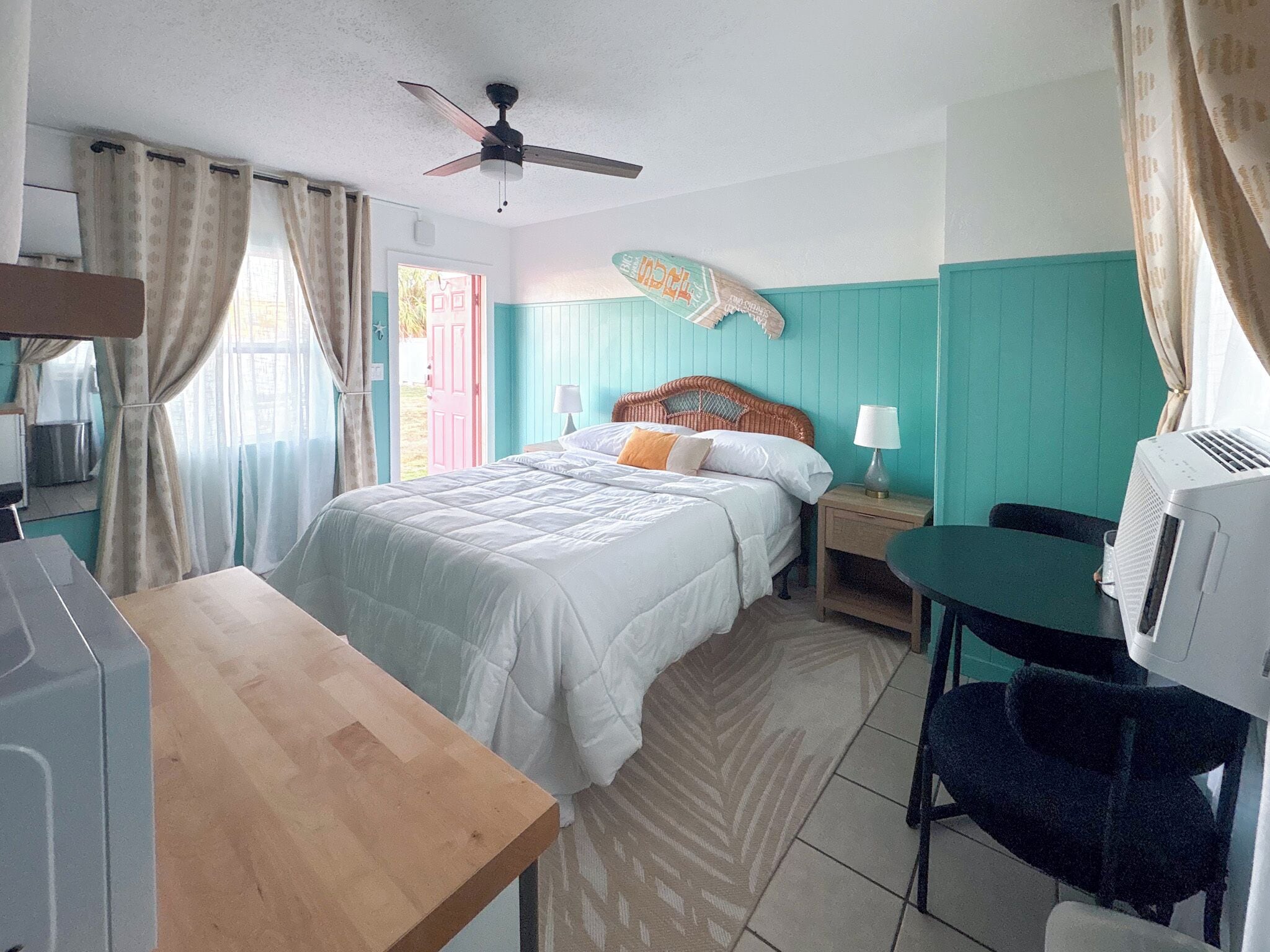 Een slaapkamer met een bed, een bureau, een stoel, een plafondventilator en een raam met gordijnen.