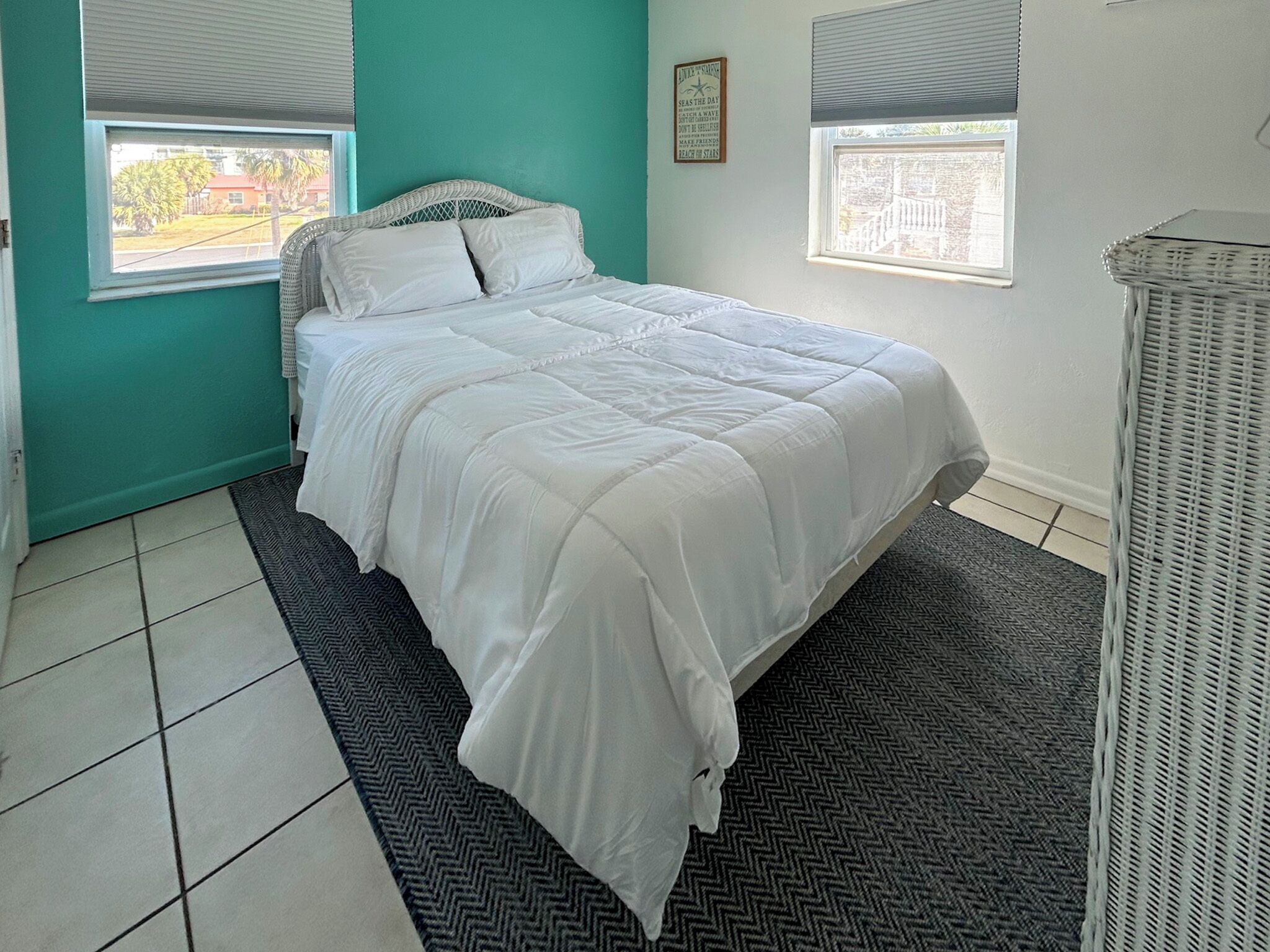 A bedroom with a bed, two windows, a wall clock, and a small wicker chair.